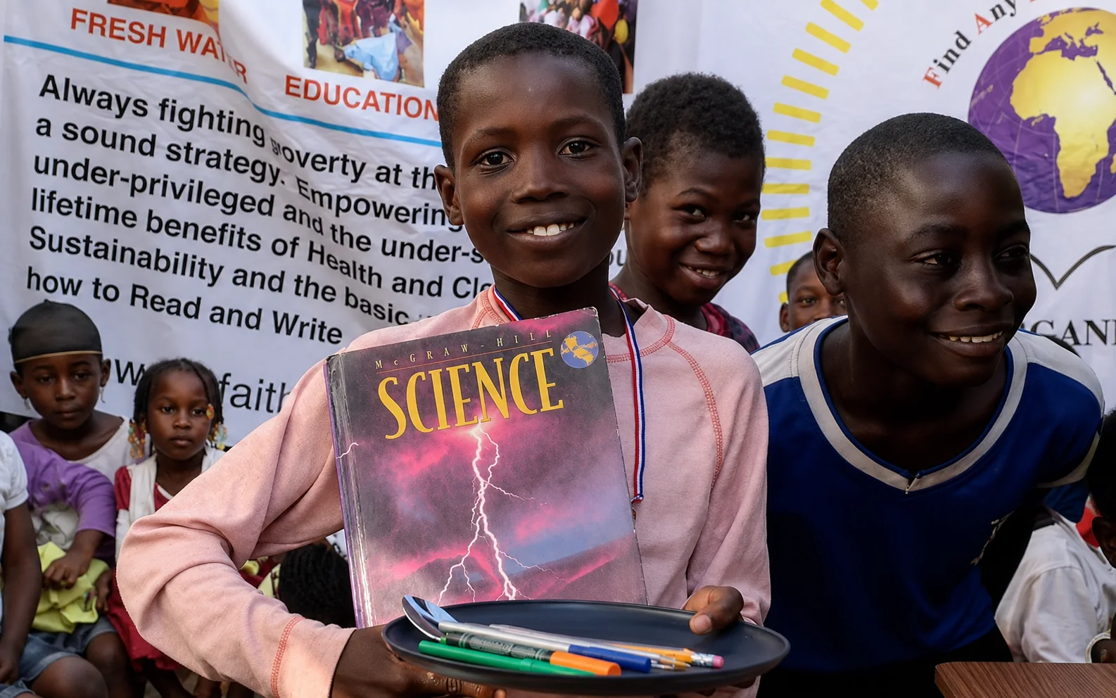 Smiling child holding a science book and school supplies during a F.A.I.T.H. Worldwide education activity